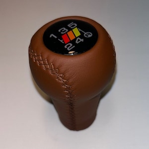 Può includere: Pomello del cambio in pelle marrone con una parte superiore circolare nera che presenta uno schema di cambio a 5 marce bianco con accenti rossi, arancioni e gialli.