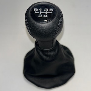 May include: A black leather gear shift knob with a matching black leather boot. The top of the knob features a glossy circular insert displaying a white manual transmission shift pattern with R, 1, 2, 3, 4, and 5 gear positions clearly printed.