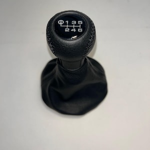 May include: A black leather gear shift knob with a matching black leather boot. The top of the knob features a white gear pattern diagram showing six forward gears and a reverse gear labeled R, 1, 2, 3, 4, 5, and 6 in a standard layout.