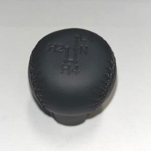 May include: A black gear shift knob with stitched detailing. The top of the knob has the letters and numbers "H2", "L", "N", and "H4" embossed on the surface. The knob is a solid, matte black colour.