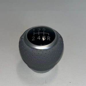 Può includere: Un pomello del cambio grigio, perforato, con una parte superiore nera che mostra lo schema delle marce: 1, 3, 5, 2, 4, 6 e R. Il pomello ha una finitura argentata ed è per un cambio manuale.