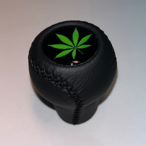 Könnte beinhalten: Schwarzer Leder-Schalthebelknauf mit einem grünen Marihuana-Blatt-Design auf der Oberseite.