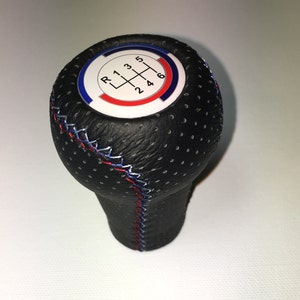 Può includere: Pomello del cambio in pelle nera con uno schema a 6 marce bianco, rosso e blu. Il pomello ha una superficie perforata e cuciture blu e rosse.