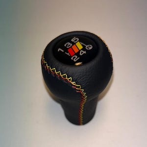May include: Black leather gear shift knob with yellow and red stitching. The knob has a white face with black numbers 1, 2, 3, 4, 5, and R printed on it.