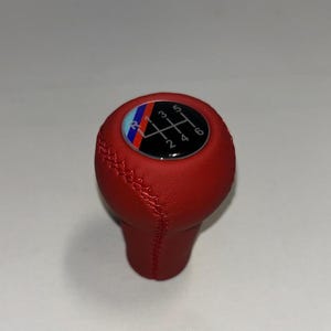 May include: A red leather gear shift knob featuring a circular top insert with a six-speed manual transmission pattern. The insert displays the numbers 1 through 6 and R, accented by a diagonal blue, purple, and red stripe design.