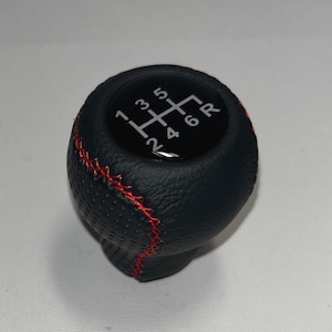 Può includere: Pomello del cambio nero con cuciture rosse. La parte superiore mostra uno schema del cambio con i numeri da 1 a 6 e R per la retromarcia. Il pomello è realizzato in materiale strutturato.