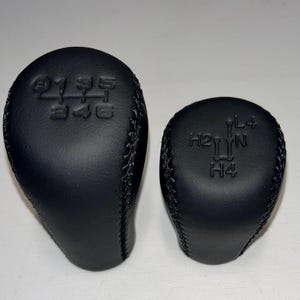 May include: Two black leather gear shift knobs. The larger knob has the numbers and layout for a standard transmission. The smaller knob has the layout for a 4-wheel drive transfer case. Both knobs have stitched seams.