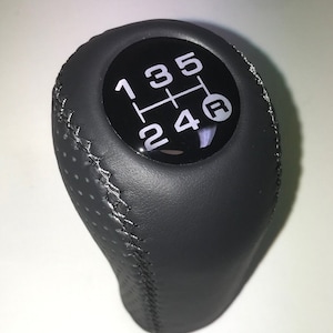 Może przedstawiać: Czarny skórzany gałka zmiany biegów z czarno-białym schematem 5-biegów i białym "R" dla biegu wstecznego.