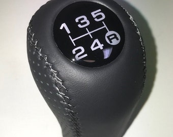 Può includere: Pomello del cambio in pelle nera con uno schema a 5 marce in bianco e nero e una "R" bianca per la retromarcia.