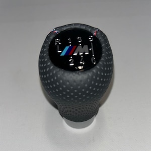May include: A dark gray, textured shift knob with a black top. The top features the BMW M logo in blue, red, and white, along with the gear shift pattern. The knob has a perforated design.