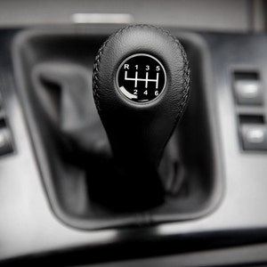 Può includere: Un pomello del cambio in pelle nera con un motivo a ingranaggio argentato e i numeri da 1 a 6 e R per la retromarcia.