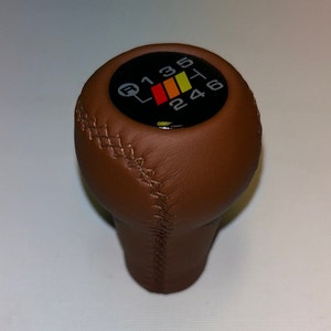 Könnte beinhalten: Brauner Lederschaltknauf mit einer schwarzen runden Oberseite, die ein weiß-gelbes Schaltmuster mit den Zahlen 1, 2, 3, 4, 5 und 6 zeigt.