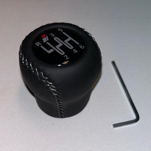 Puede incluir: Pomo de cambio de marchas de cuero negro con costuras blancas y un patrón de engranaje plateado con los números 1, 2, 3, 4, 5 y 6. La letra R también está en el pomo.