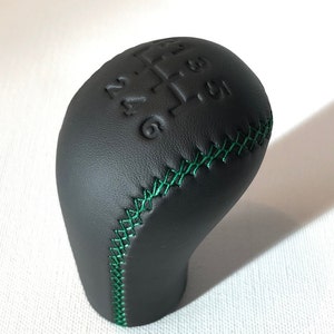 Könnte beinhalten: Schwarzer Lederschaltknauf mit grünen Nähten. Der Knauf hat geprägte Zahlen 1, 2, 3, 4, 5 und 6.