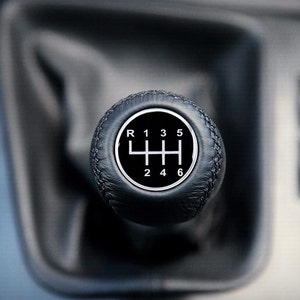Op de afbeelding: Een zwarte leren versnellingspookknop met witte cijfers en een versnellingschema. De cijfers op de knop zijn R, 1, 2, 3, 4, 5 en 6.