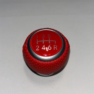 Könnte beinhalten: Roter Schalthebelknauf mit silbernem Zierring. Die Oberseite hat einen roten Kreis mit dem Gangschaltschema in Weiß: 1, 3, 5, H, 2, 4, 6 und R. Der Knauf hat eine strukturierte Oberfläche.