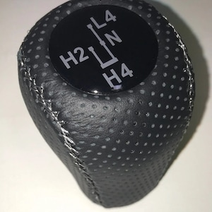 Può includere: Pomello del cambio in pelle nera con un adesivo circolare nero e bianco che dice "H2 L4 N H4".