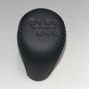 Peut inclure: Un pommeau de levier de vitesses en cuir noir avec une couture. Le dessus du pommeau porte en relief le schéma des vitesses : R, 1, 3, 5, 2, 4, 6.