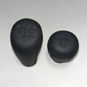 May include: Two black leather gear shift knobs. One is elongated with numbers 1 to 6, and the other is round with H2, N, and H4 markings. Both have stitched seams.