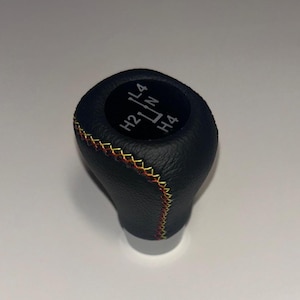 Op de afbeelding: Een zwarte, getextureerde versnellingspookknop met een ronde inleg aan de bovenzijde waarop in witte tekst het schakelpatroon L4, N, H2 en H4 staat. De knop is voorzien van decoratieve kruissteken in een verloop van geel, oranje en rood garen.