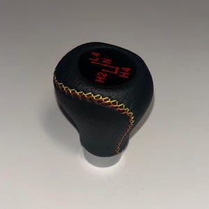Op de afbeelding: Een zwarte, getextureerde versnellingspookknop met een ronde inzet aan de bovenzijde voorzien van rode L4, N, H2 en H4 versnellingsmarkeringen. De knop is afgewerkt met decoratieve kruissteken in afwisselend geel en oranje garen.