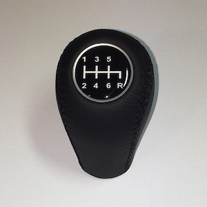 Puede incluir: Pomo de cambio de marchas de cuero negro con un anillo plateado y números blancos 1, 2, 3, 4, 5, 6 y R para la marcha atrás.