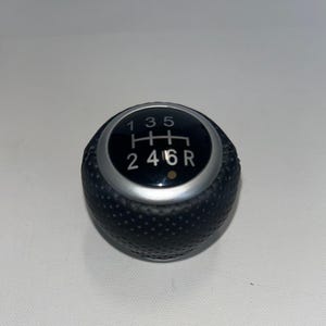 May include: A black gear stick knob with a silver trim. The top features a black circle with white numbers and letters indicating gear positions: 1, 3, 5, H, 2, 4, 6, and R. The knob has a textured surface.