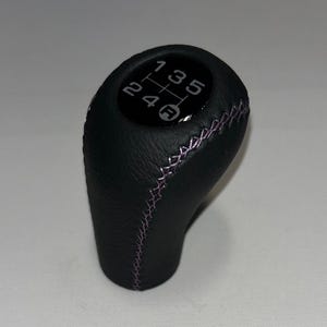 Op de afbeelding: Zwarte lederen versnellingspookknop met paarse stiksels. De bovenkant van de knop toont het schakelpatroon in witte tekst: 1, 2, 3, 4, 5 en R (achteruit).