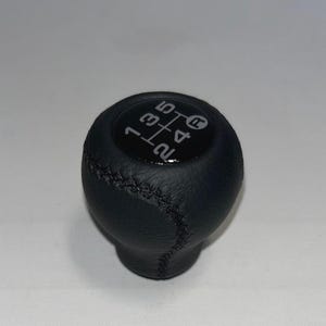 May include: A black, leather-wrapped shift knob with white gear shift pattern markings. The shift pattern includes numbers 1 through 5, plus reverse. The knob has a stitched seam and a rounded shape.