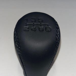 May include: A black, leather-wrapped gear shift knob. The top of the knob has the gear pattern embossed in the leather: 1, 3, 5, 2, 4, 6, and R. The knob is designed for a manual transmission.