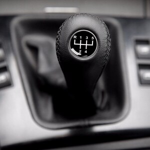 Op de afbeelding: Een zwarte leren versnellingspookknop met een zilveren tandwielpatroon en de nummers 1 tot en met 5 en de letter R.