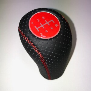 Pode incluir: Botão de câmbio de couro preto com costuras vermelhas e um círculo vermelho com números brancos 1, 2, 3, 4, 5, 6 e R.