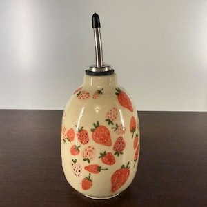 May include: A cream-colored ceramic oil dispenser with a hand-painted strawberry pattern in shades of red and pink. The bottle features a rounded body and is topped with a stainless steel pour spout with a black plastic cap.