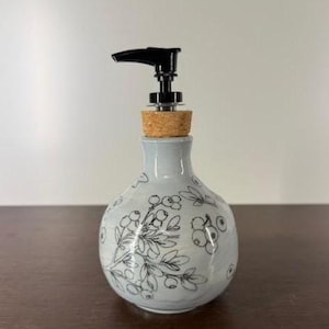 Puede incluir: Un dispensador de jabón de cerámica con un esmalte azul claro y una bomba negra. El dispensador está decorado con un diseño floral y de bayas negras. El dispensador tiene un tapón de corcho y está sobre una superficie marrón oscuro.