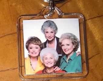 May include: A clear plastic keychain with a picture of four women. The women are all wearing different colored shirts and have gray hair. The keychain is attached to a silver metal ring.
