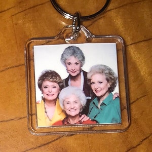 May include: A clear plastic keychain with a picture of four women. The women are all wearing different colored shirts and have gray hair. The keychain is attached to a silver metal ring.