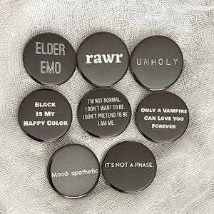 May include: Nine black button pins with white text. The pins feature phrases like "Elder Emo", "rawr", "Unholy", "Black is my happy color", "I'm not normal. I don't want to be. I don't pretend to be. I am me.", "Only a vampire can love you forever", "Mood: apathetic", and "It's not a phase."