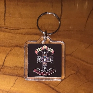 May include: Clear square keychain featuring the Guns N' Roses logo. The design includes a cross with band member portraits and the band's name in a banner. The keychain has a silver metal ring.