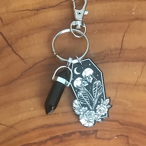 May include: Black and white enamel keychain featuring a couple of skeletons embracing in a coffin with flowers and a crescent moon. The keychain also includes a black crystal charm.