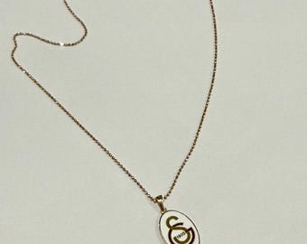 Puede incluir: Collar dorado con una cadena delicada y un colgante ovalado. El colgante presenta un fondo blanco con un emblema dorado y verde. Un accesorio elegante.