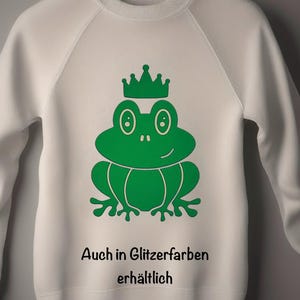 Könnte beinhalten: Weißes Sweatshirt mit einer grünen Frosch-Grafik, die eine Krone trägt. Das Frosch-Design ist mittig auf der Vorderseite platziert. Darunter steht der Text "Auch in Glitzerfarben erhältlich". Das Sweatshirt hängt an einem Holzkleiderbügel.
