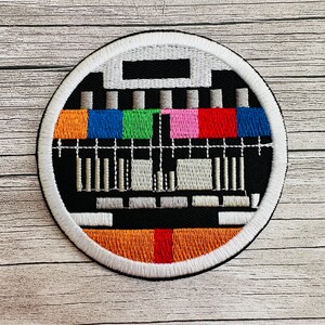 Könnte beinhalten: Ein runder gestickter Aufnäher mit weißem Rand und einem schwarz-weißen Fernseh-Testbild in der Mitte. Das Muster enthält farbige Balken in Rot, Blau, Grün, Orange und Pink.