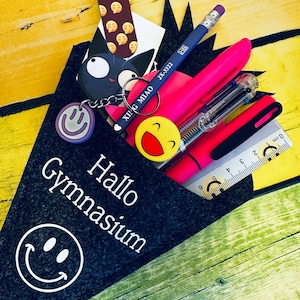 Könnte beinhalten: Ein schwarzes Filz-Federmäppchen gefüllt mit Schulmaterialien, darunter ein Bleistift, Stifte, ein Lineal und ein Lesezeichen. Das Etui trägt den Text "Hallo Gymnasium" und ein Smiley. Ein Schlüsselanhänger mit dem Text "XING MIAO" ist ebenfalls sichtbar.