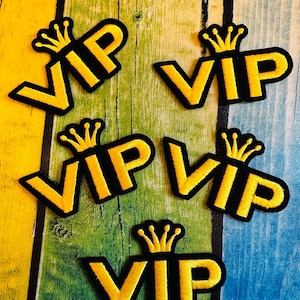 Könnte beinhalten: Eine Sammlung gestickter VIP-Patches. Jeder Patch zeigt die Buchstaben "VIP" in Gelb, umrandet von Schwarz und mit einer goldenen Krone versehen. Die Patches sind auf einer farbenfrohen, strukturierten Holzoberfläche angeordnet.