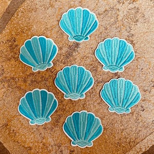 Könnte beinhalten: Sieben gestickte Aufnäher in Form von türkisfarbenen Muscheln mit weißen Rändern. Die Aufnäher sind auf einer hellbraunen Oberfläche angeordnet. Ideal zum Aufnähen auf Kleidung oder Accessoires.