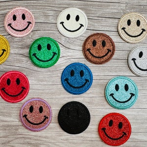 Könnte beinhalten: Zwölf verschiedene Smiley-Patches mit Glitzer und verschiedenen Farben, darunter Rot, Blau, Grün, Gold, Silber und Schwarz.