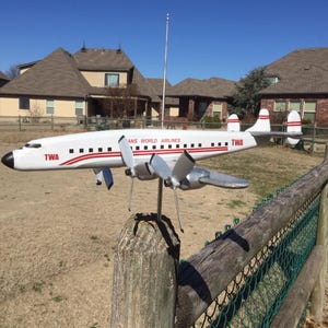 May include: A white and red model airplane with "TWA" and "Trans World Airlines" printed on the side, mounted on a wooden post with a metal fence in the background.