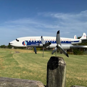 May include: A model airplane featuring a white fuselage with a blue stripe and the text FLY EASTERN AIR LINES. The aircraft has four silver propellers, a circular logo on the nose, and a small American flag decal on the tail fin assembly.