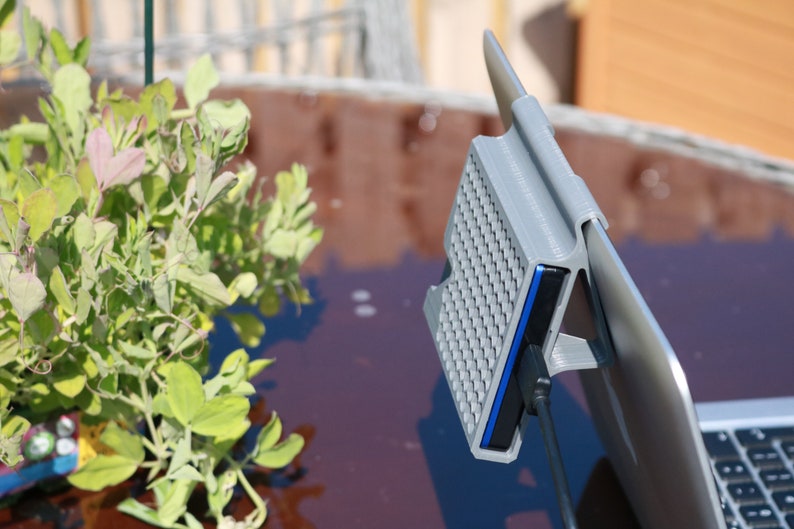 May include: A gray 3D printed hard drive enclosure with a blue USB cable attached to a laptop. The enclosure has a textured surface and is attached to the laptop with a clip.