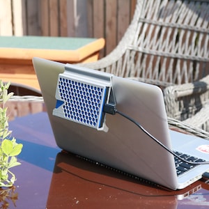 May include: A silver laptop computer with a blue and silver cooling fan attached to the back. The laptop is sitting on a wooden table with a green plant in a pot to the left. The laptop is plugged into an outlet with a black cord.
